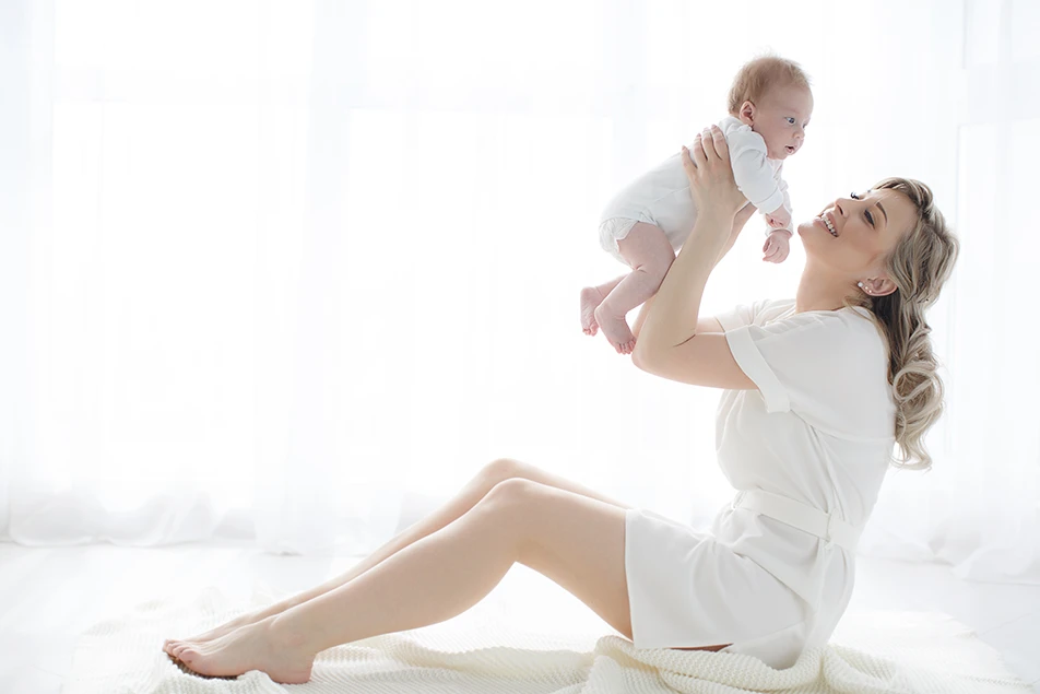 smiling mother holding her baby in a bright nursery, symbolising life after childbirth and the journey to restore her post-pregnancy body.